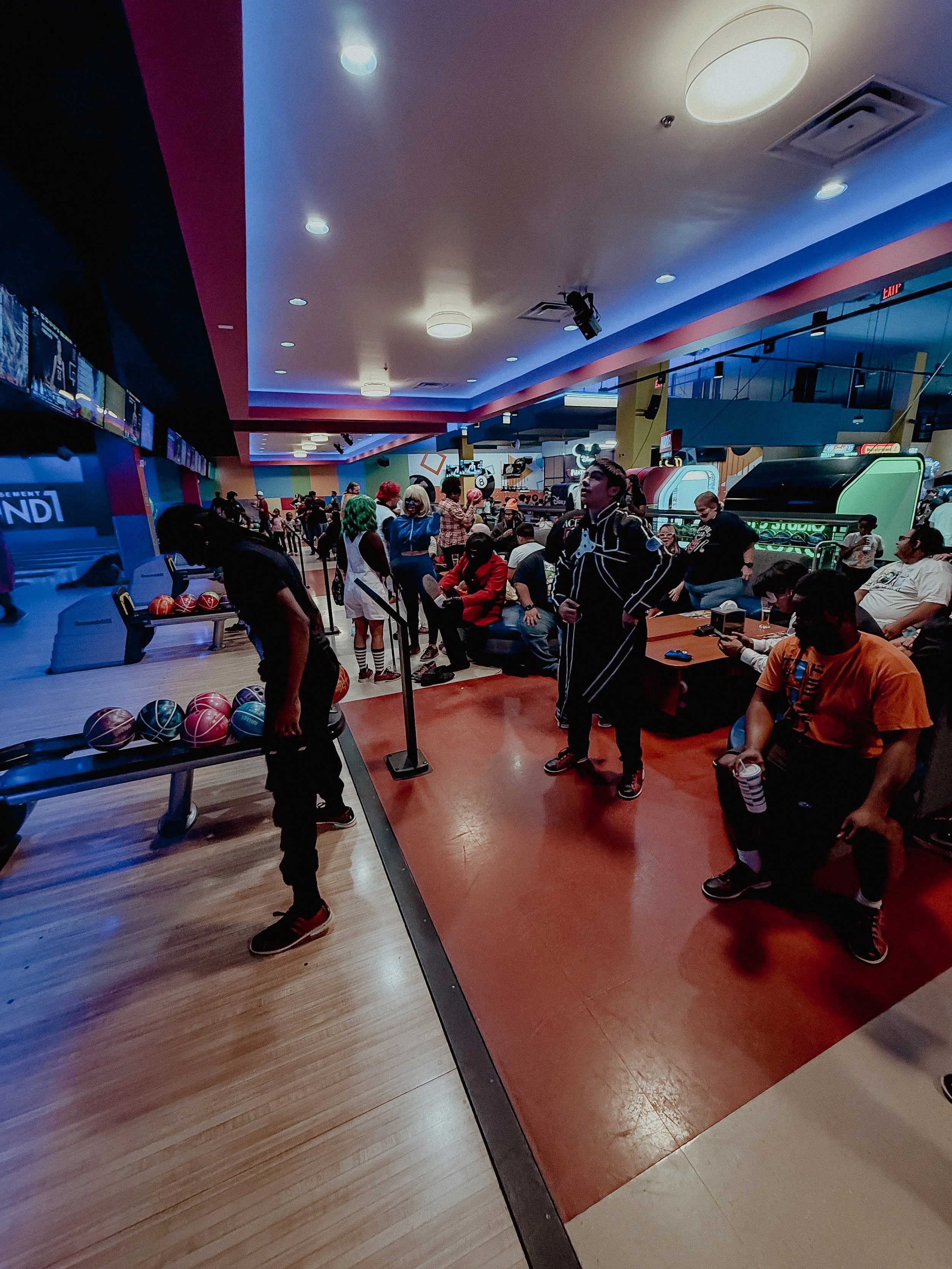People at a bowling alley, some sitting and some standing, with bowling balls on racks and screens above lanes, with colorful lighting and arcade games in the background.
