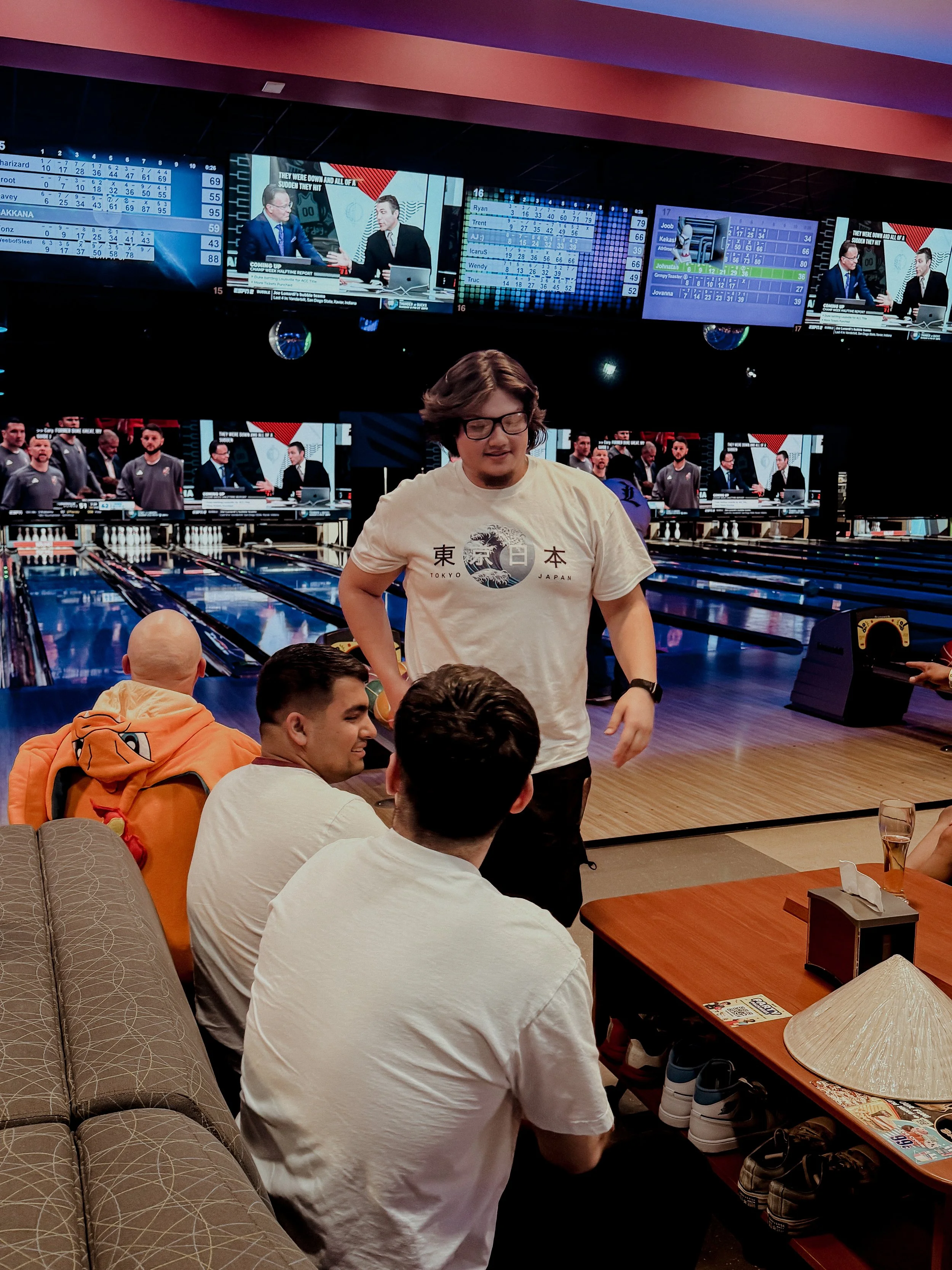 People in a bowling alley, with one person standing and three seated. The background shows large screens displaying scores and a broadcast, with several others playing or watching in the background.