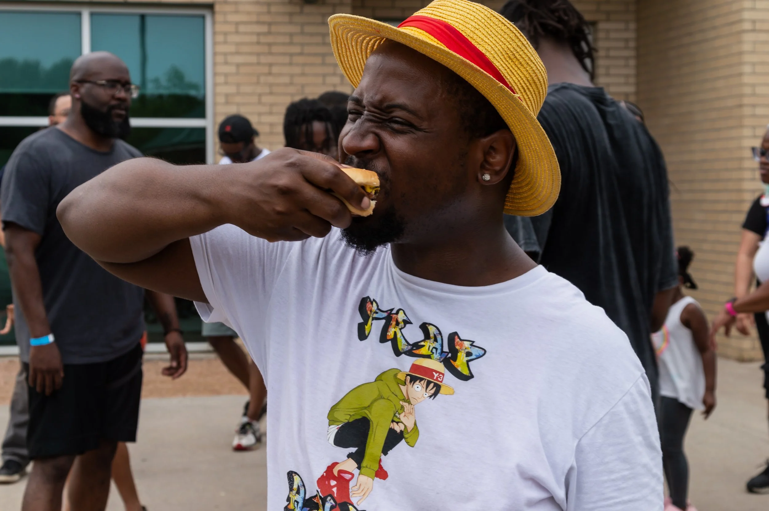 A man wearing a yellow Luffy hat with a red band, white T-shirt with a cartoon character, is eating a sandwich at an outdoor gathering, with people standing in the background.
