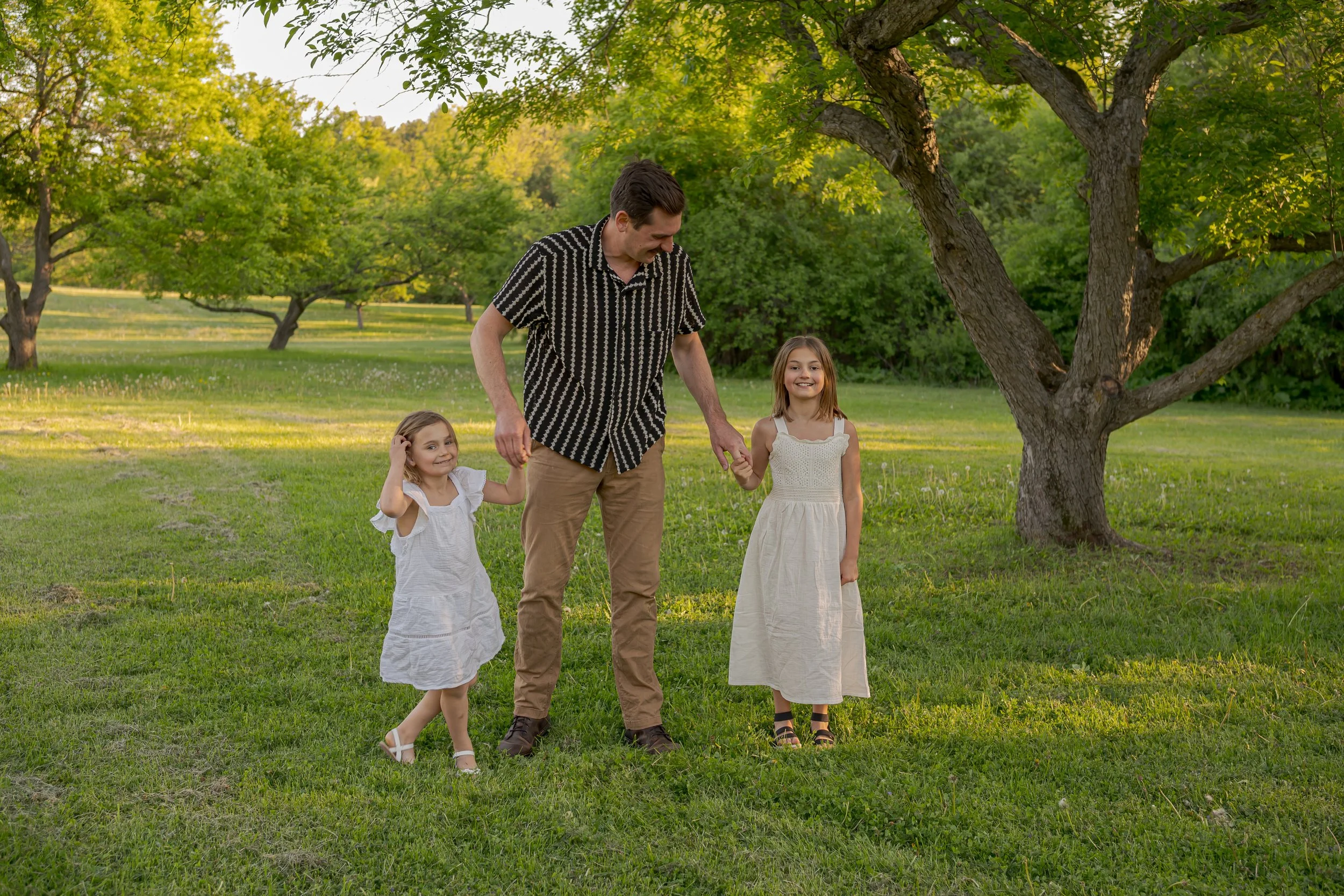 A father walking with his two young daughters in a park with green grass and trees during daylight.