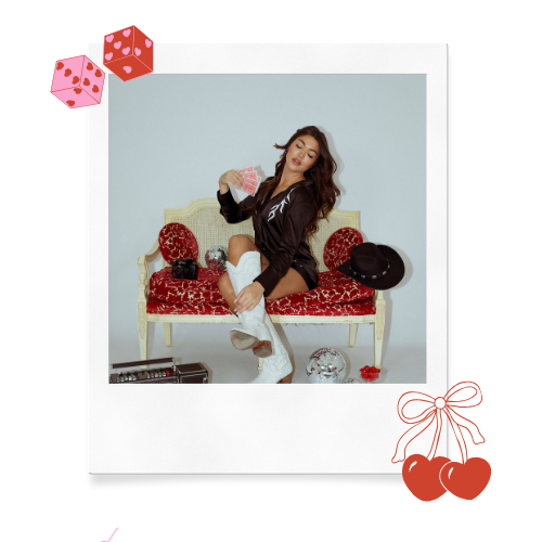 A woman sitting on a vintage red and white ornate loveseat holding a stack of dollar bills. She is wearing a black jacket, white knee-high socks, and beige shoes. There are black handbags and a black hat on the loveseat. A small glass bowl, a small guitar, and a red rose are on the floor. The background is plain white with decorative Valentine's Day-themed red and pink gift box graphics and hearts.