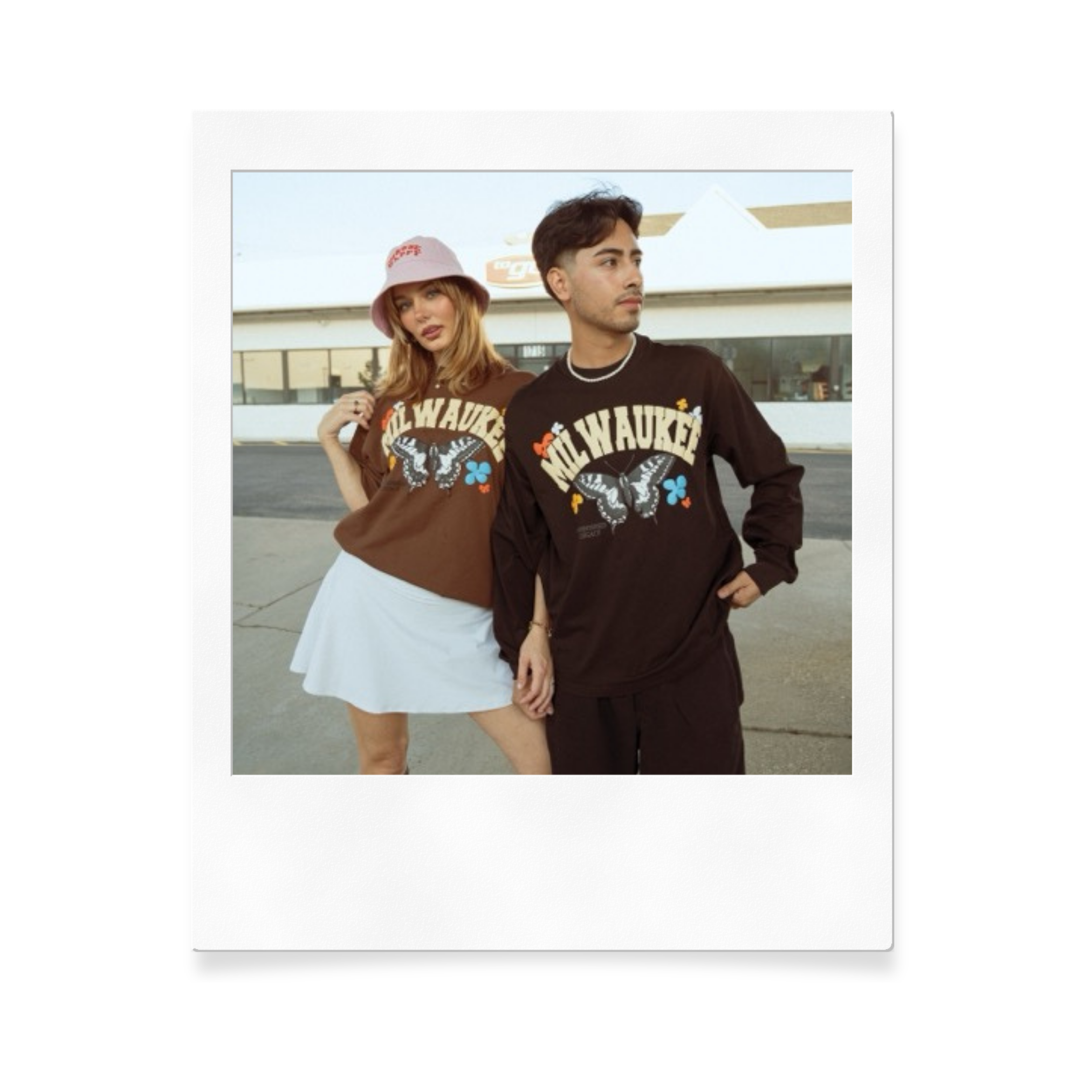 Two young adults, a woman and a man, standing outdoors in front of a modern building, wearing matching brown and black Milwaukee graphic sweatshirts with butterfly and flower designs.