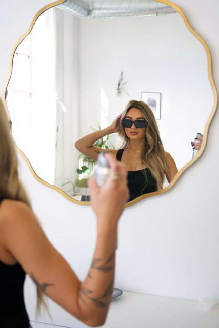 Woman taking a photo of herself in a mirror, wearing sunglasses and a black top, in a bright room with a potted plant and framed picture on the wall.