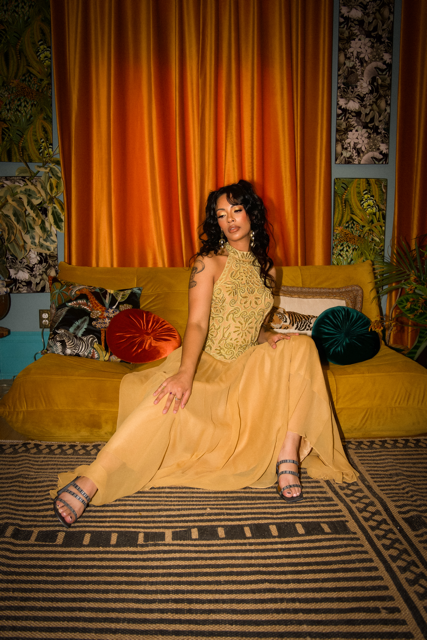 A woman wearing a yellow dress sitting on a yellow couch in a colorful room with orange curtains, patterned pillows, and vibrant wall art.