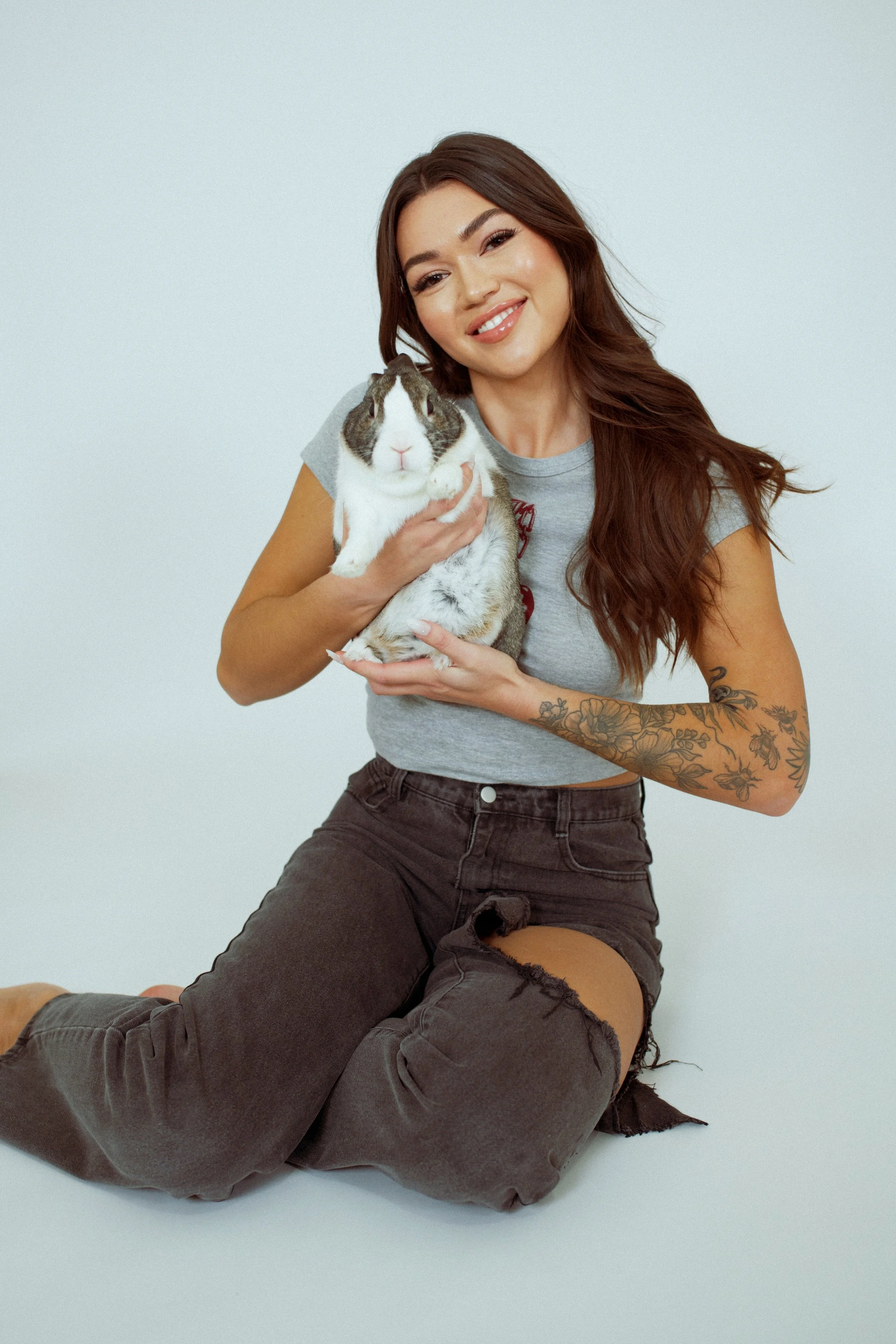 A woman with long brown hair, tattoos on her arm, smiling, holding a guinea pig, sitting on the floor against a plain white background.