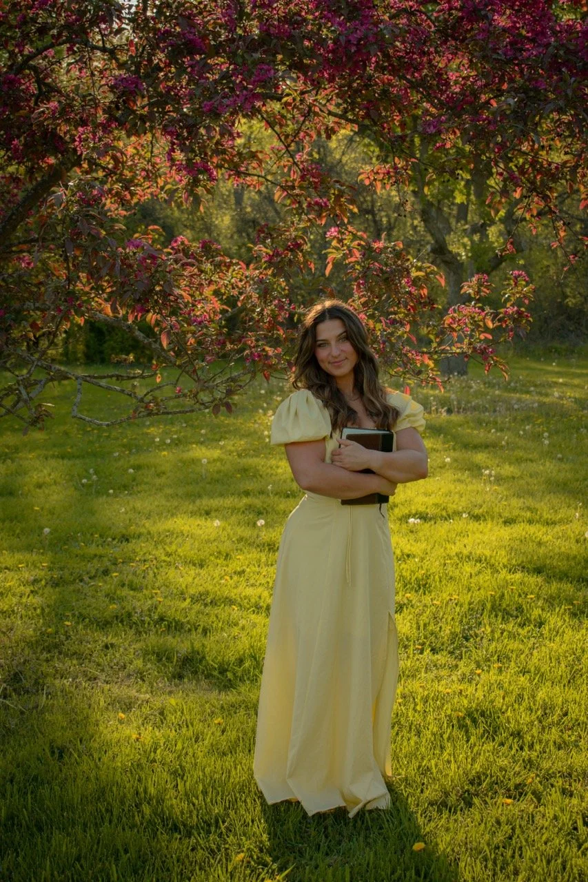 A woman in a yellow dress holding books stands on a grassy field under a flowering tree with pink blossoms, during sunset.