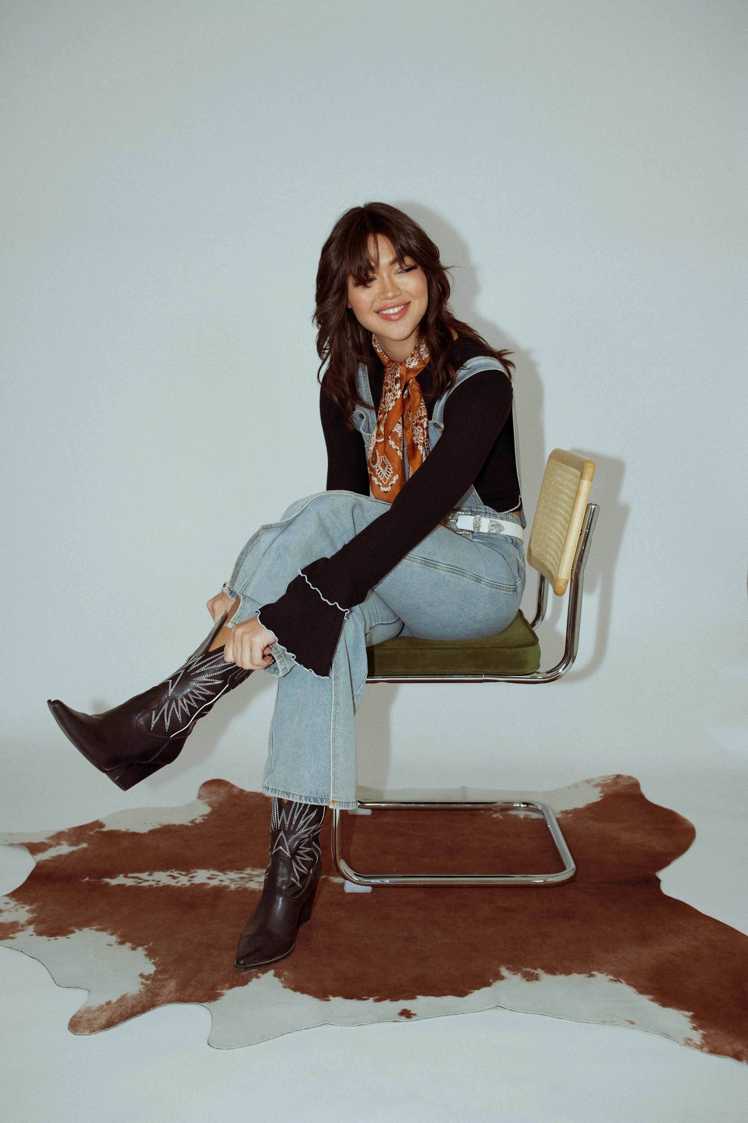 A woman sitting on a modern office chair on a cowhide rug, wearing cowboy boots, jeans, a black top, and a patterned orange bandana, smiling at the camera.