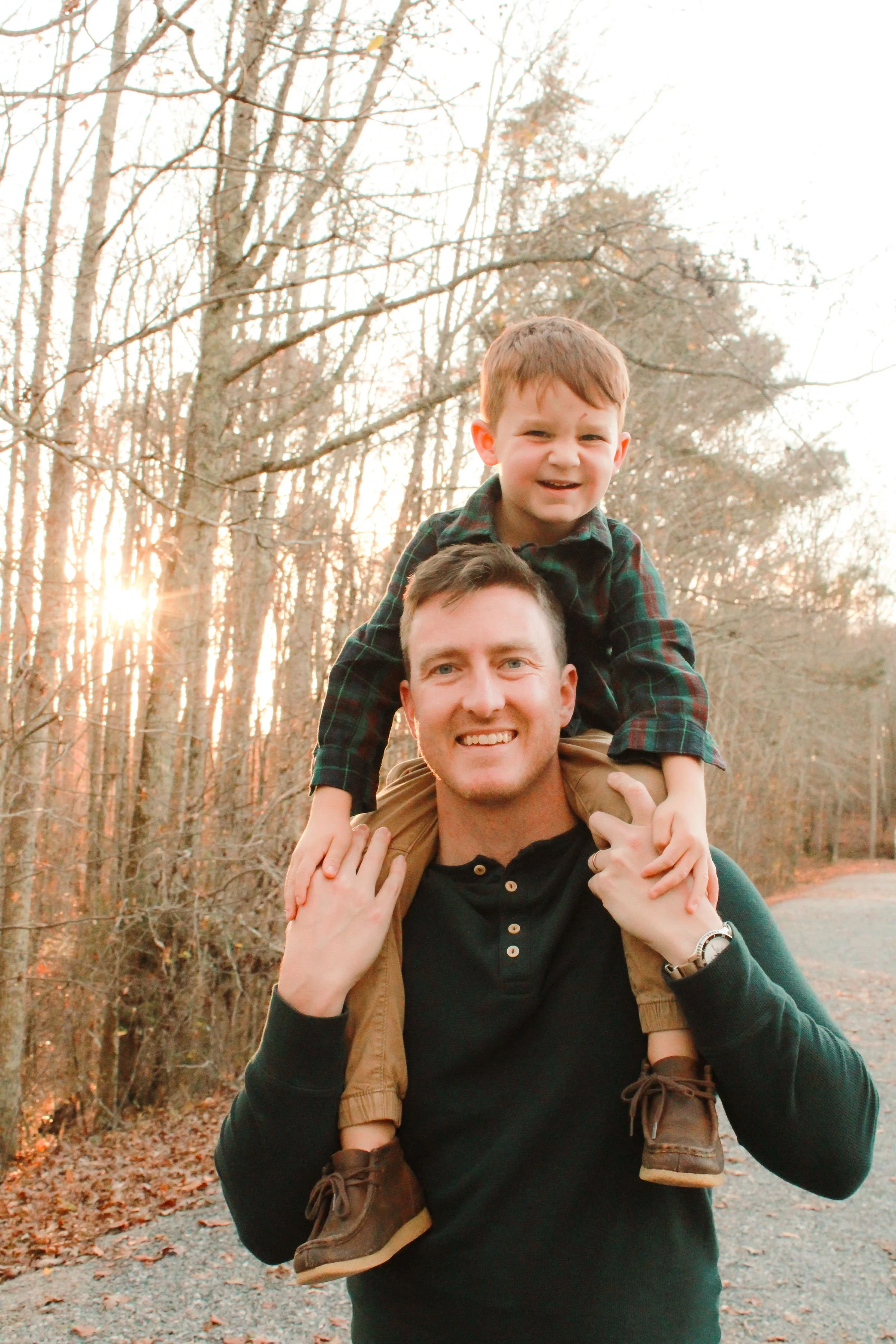 A man carrying a young boy on his shoulders in a wooded area during sunset, both smiling and enjoying the outdoors.