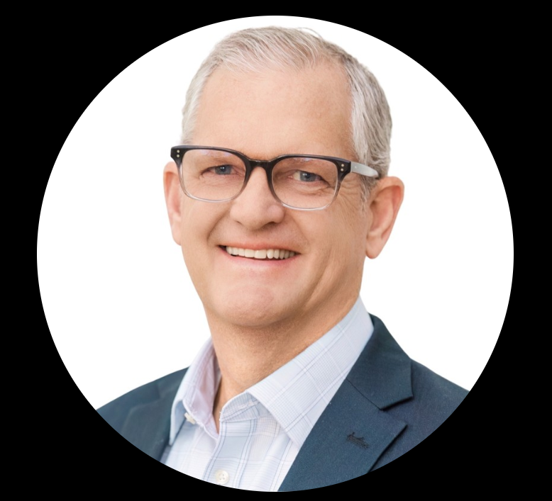 A smiling middle-aged man with gray hair and glasses, wearing a business suit and a white collared shirt, posing against a white background with a black border.