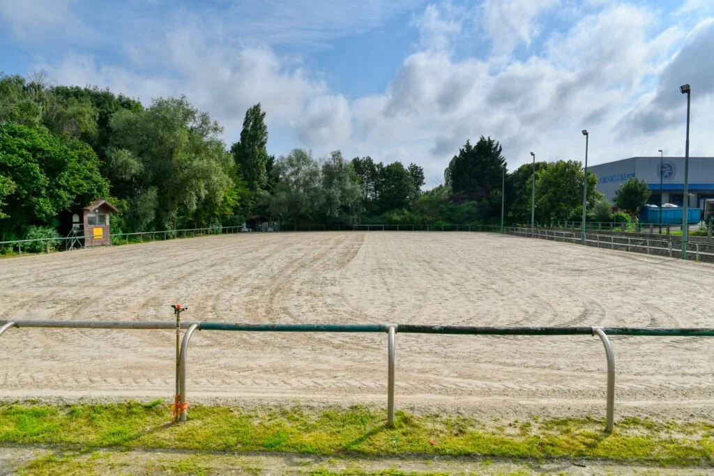 Un terrain de sport en sable entouré de barrières métalliques, avec des arbres verts en arrière-plan et un bâtiment à droite, sous un ciel partiellement nuageux.