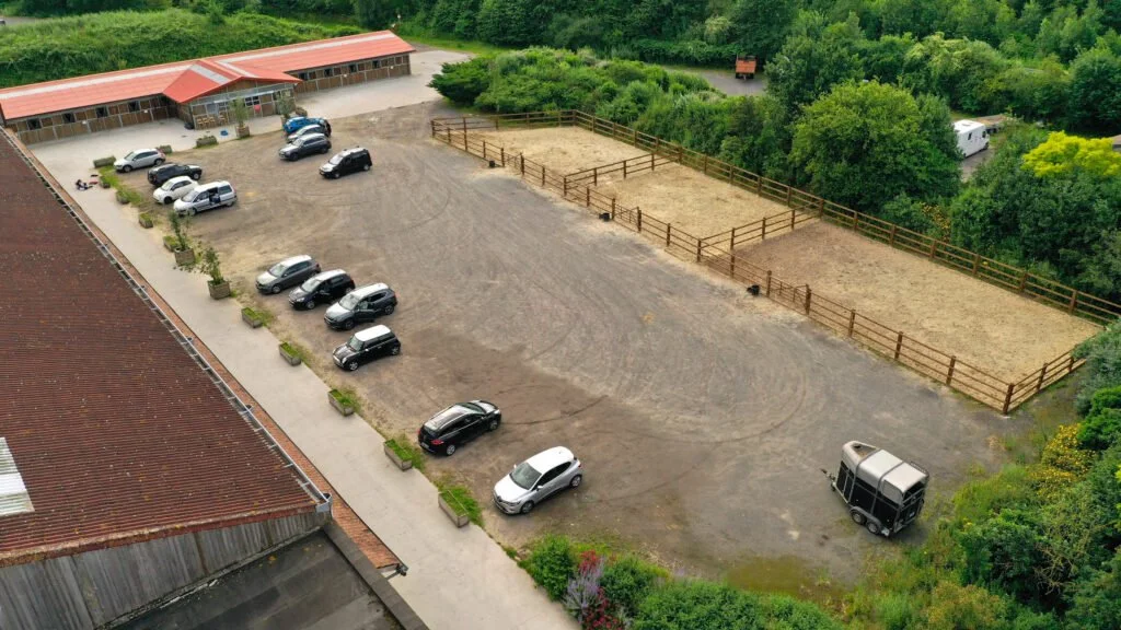 Une vue aérienne d'un centre équestre avec deux zones de paddocks clôturés, un bâtiment, plusieurs voitures garées et un environnement verdoyant.