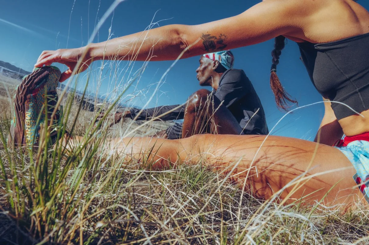 A person stretching outdoors in a grassy field, with another person sitting nearby, both under a clear blue sky.
