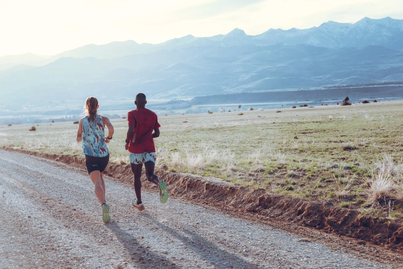 Two people running on a gravel road through a rural landscape with mountains in the background during daytime.