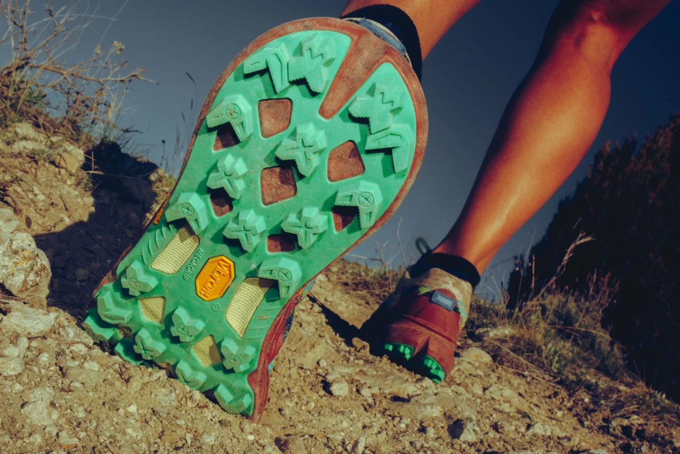 Close-up of a person hiking on rocky terrain, showing a detailed view of the sole of their hiking shoe with green and red colors, positioned on an uneven dirt trail in an outdoor setting with dry bushes and a dark sky.