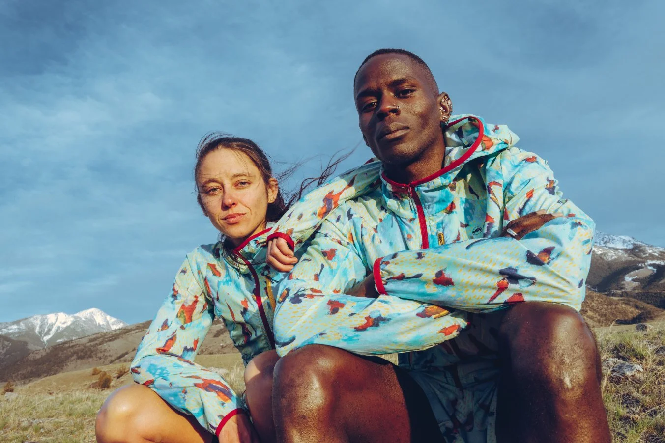 Two people in colorful jackets outdoors with mountains and snow in the background.