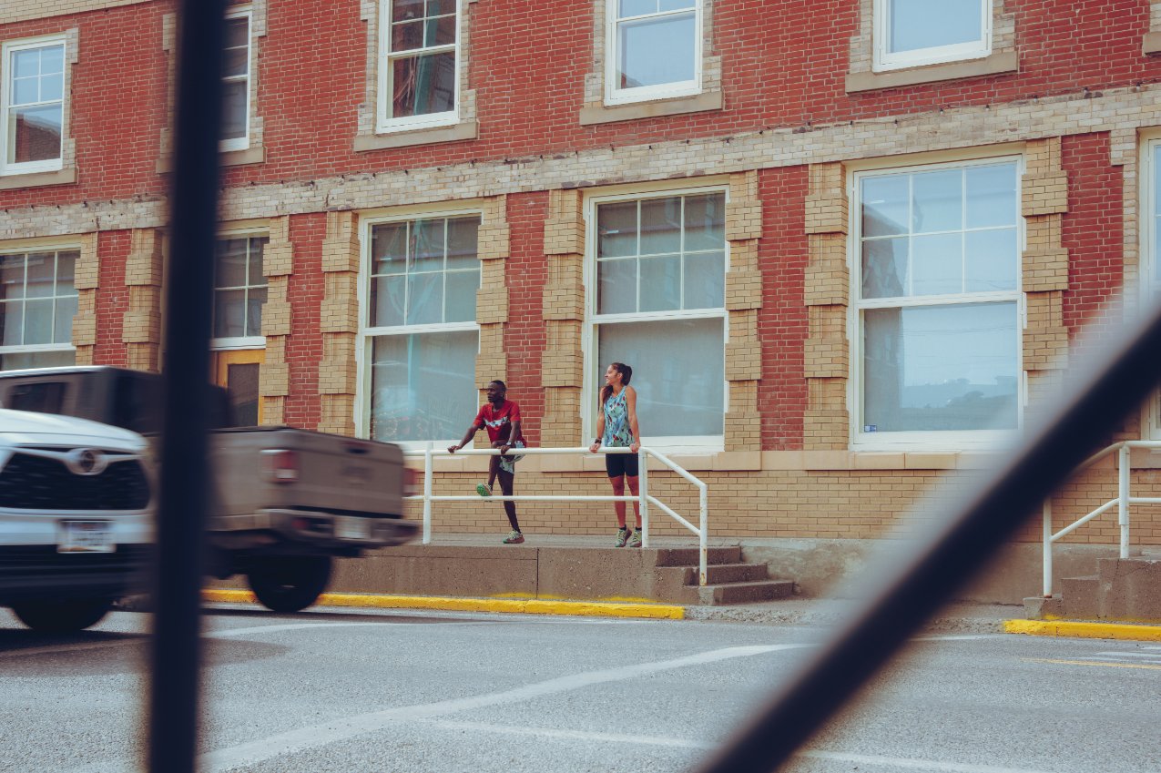 Two women walking on a sidewalk in front of a brick building with large windows, seen through a fence with a blurred vehicle passing by in the foreground.