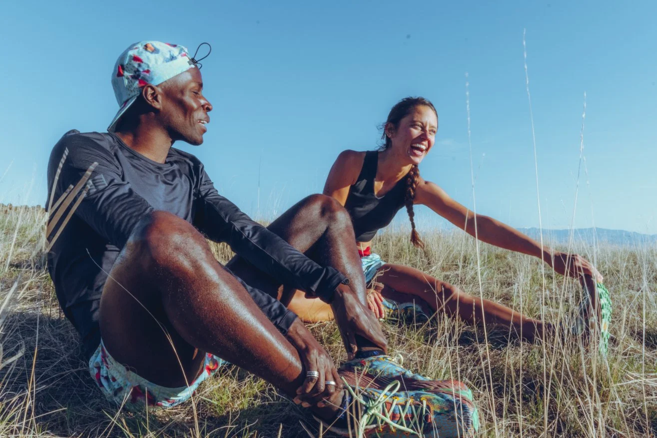 Two people sitting on grassy dunes, laughing and enjoying outdoor activity on a sunny day.