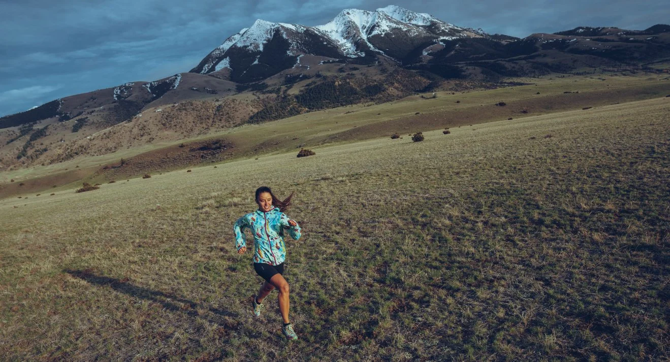 A woman runs on a grassy field with mountains in the background, some with snow, and a partly cloudy sky.