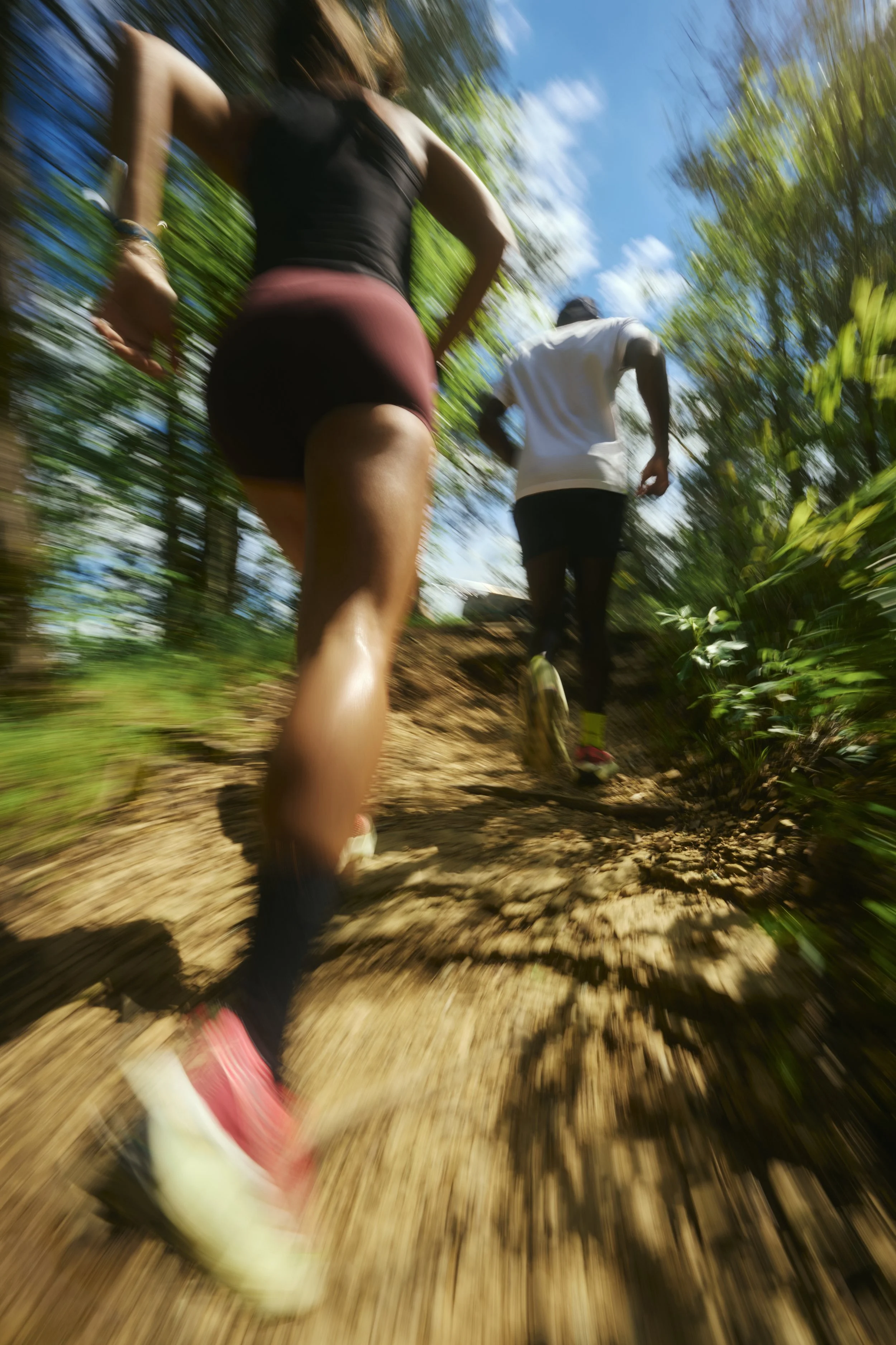 Two people running on a dirt trail through a forested area under a bright blue sky.