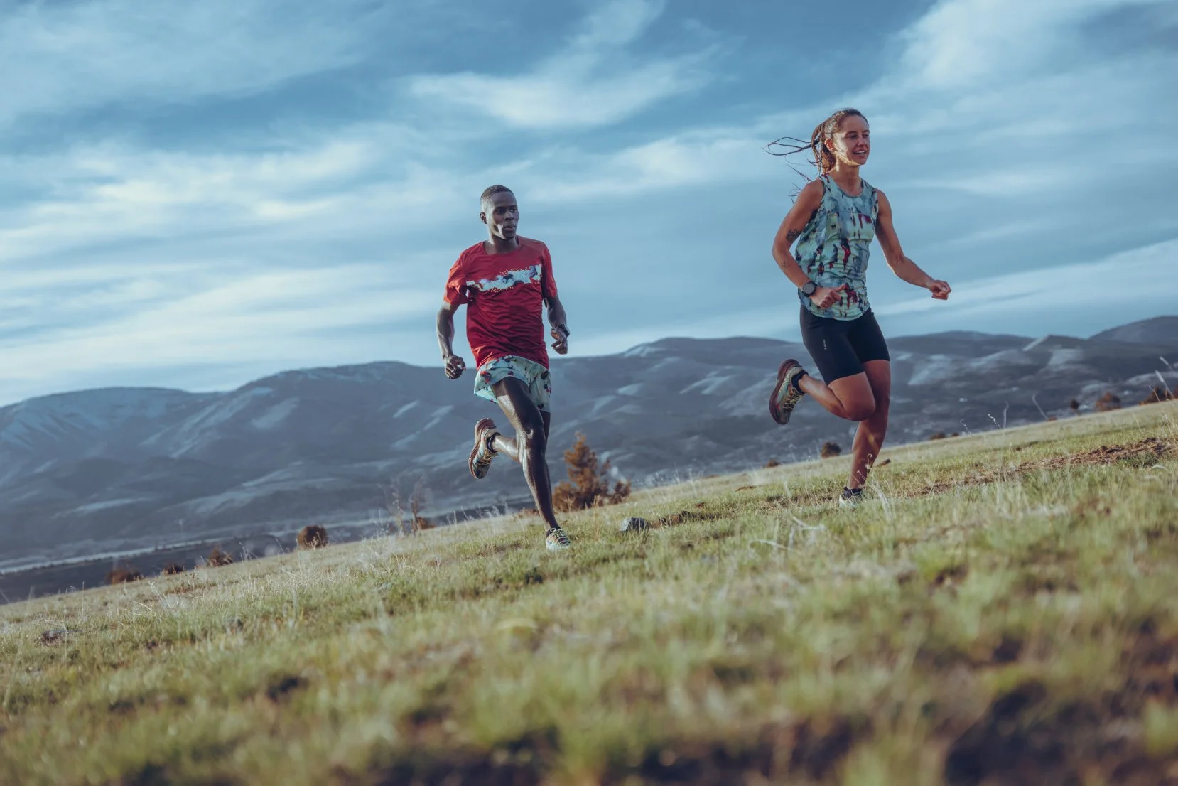 Two people running outdoors on a grassy hill with mountains and a cloudy sky in the background.