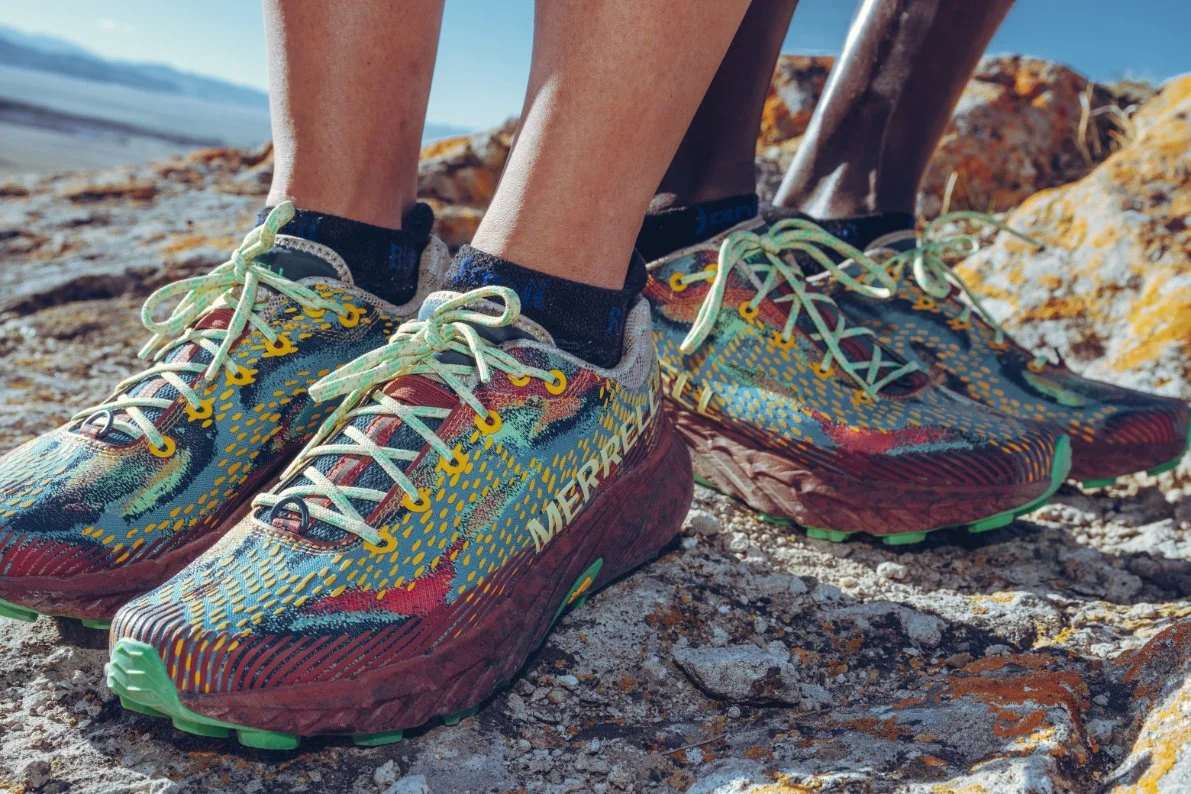 Close-up of colorful Merrell trail running shoes on rocky terrain, with two individuals in the background outdoors.
