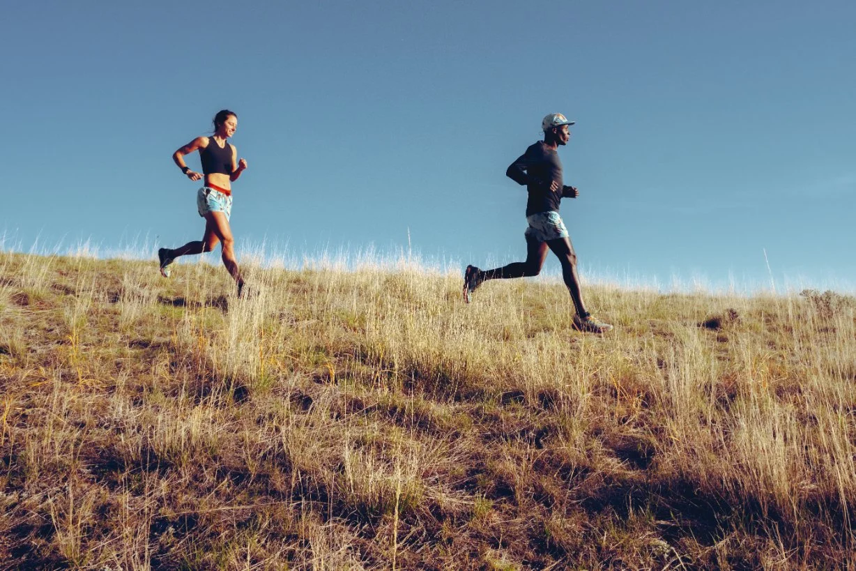 Two people trail running uphill on a grassy hill against a clear blue sky.