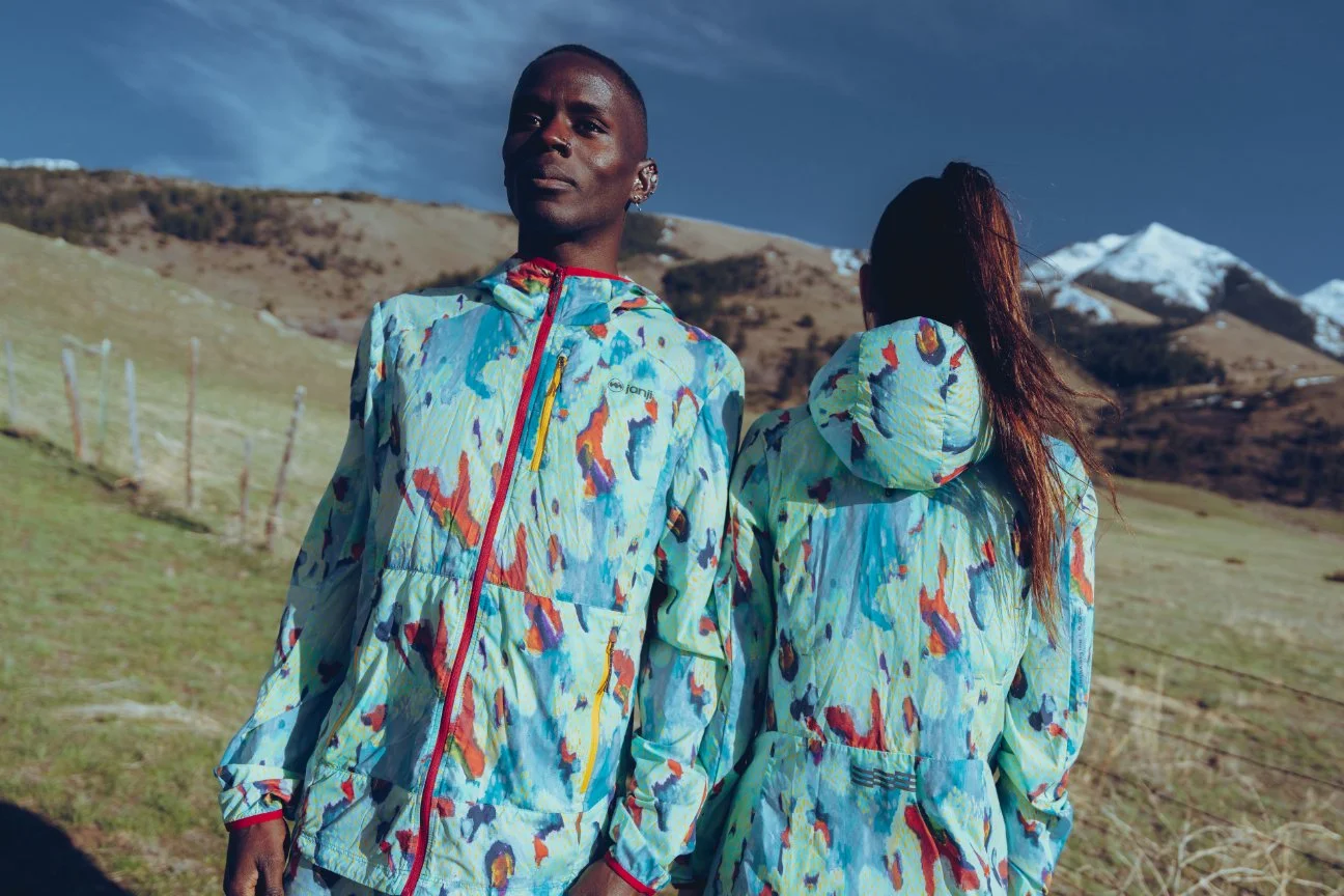 Man and woman standing outdoors on a hillside, wearing matching colorful abstract patterned jackets, mountains in background.