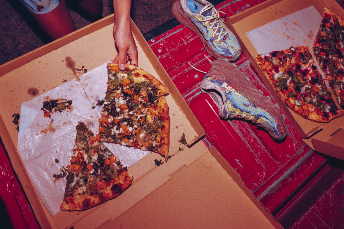 A partially eaten pizza in a cardboard box with a person's hand holding a slice, on a red wooden surface with sneakers and another pizza box in the background.