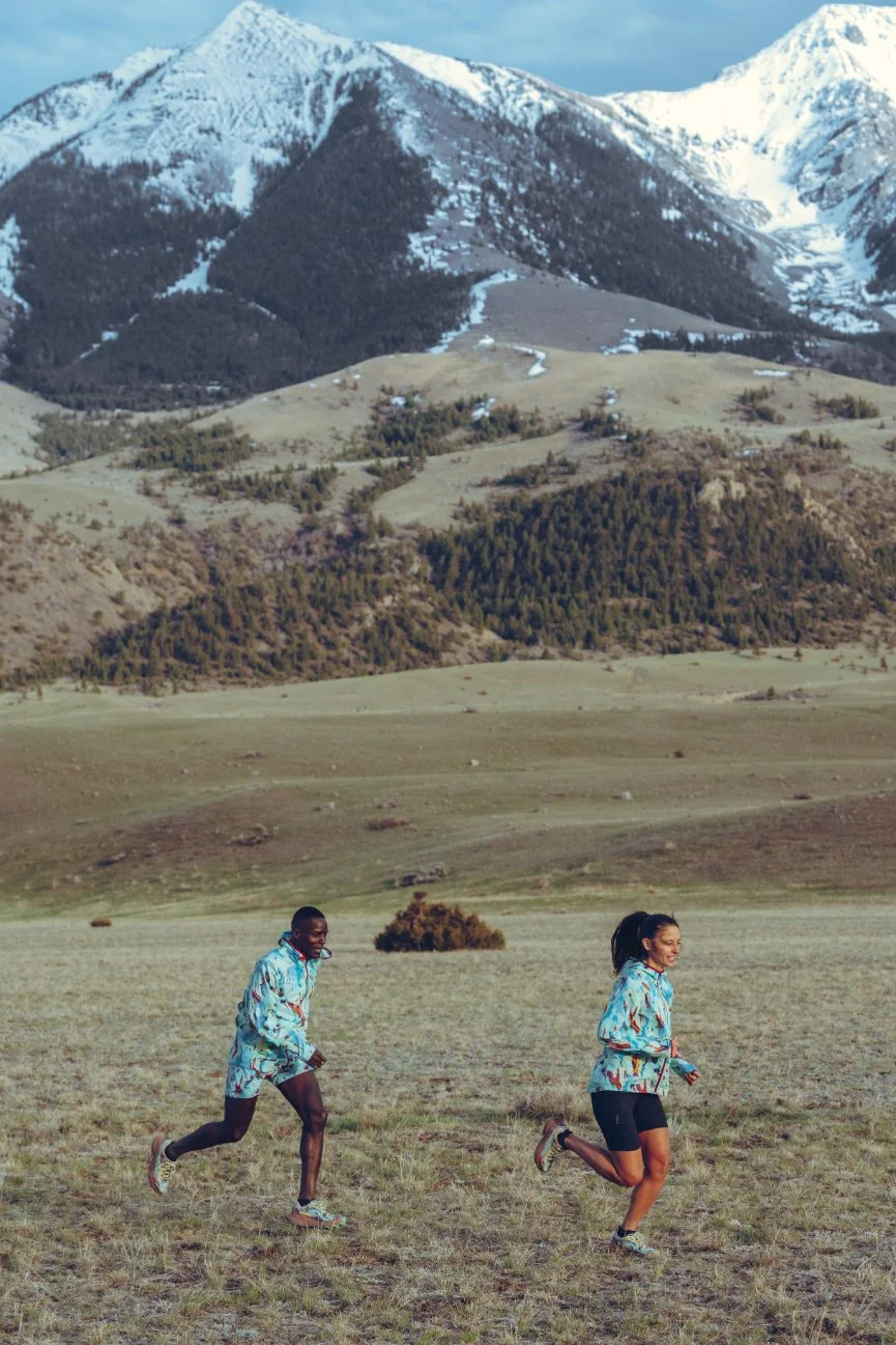Two people running across a grassy field with mountains in the background, snow-capped peaks and rolling hills.