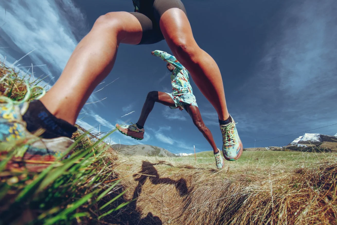 A runner in colorful athleticwear jumping over a trench in a grassy landscape with blue sky and mountains in the background, captured from a low-angle perspective.