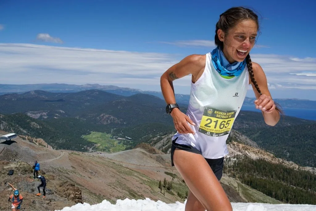 Female runner participating in a mountain race, smiling and wearing a race bib, with scenic mountain landscape background.