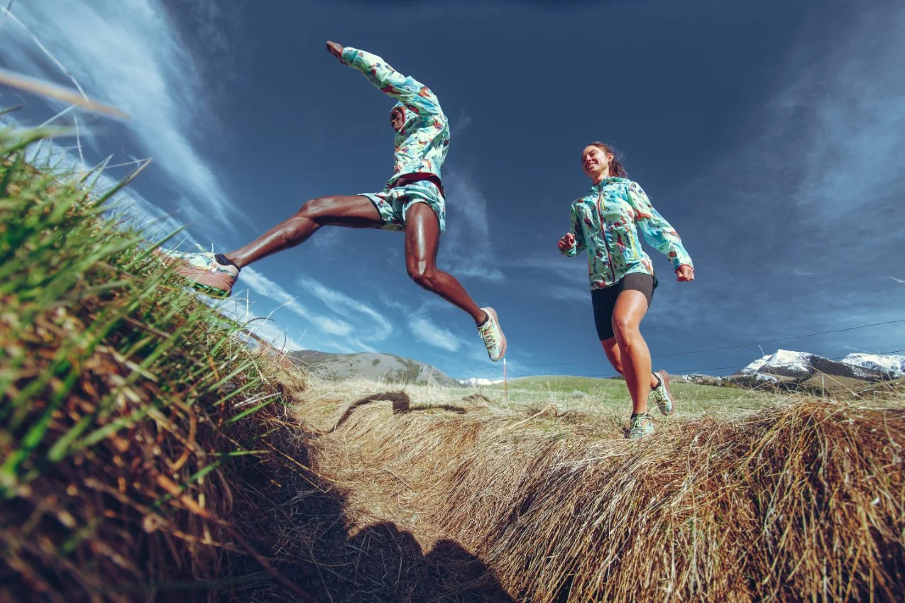 Two runners in colorful athletic wear running across a grassy field outdoors under a blue sky with clouds.