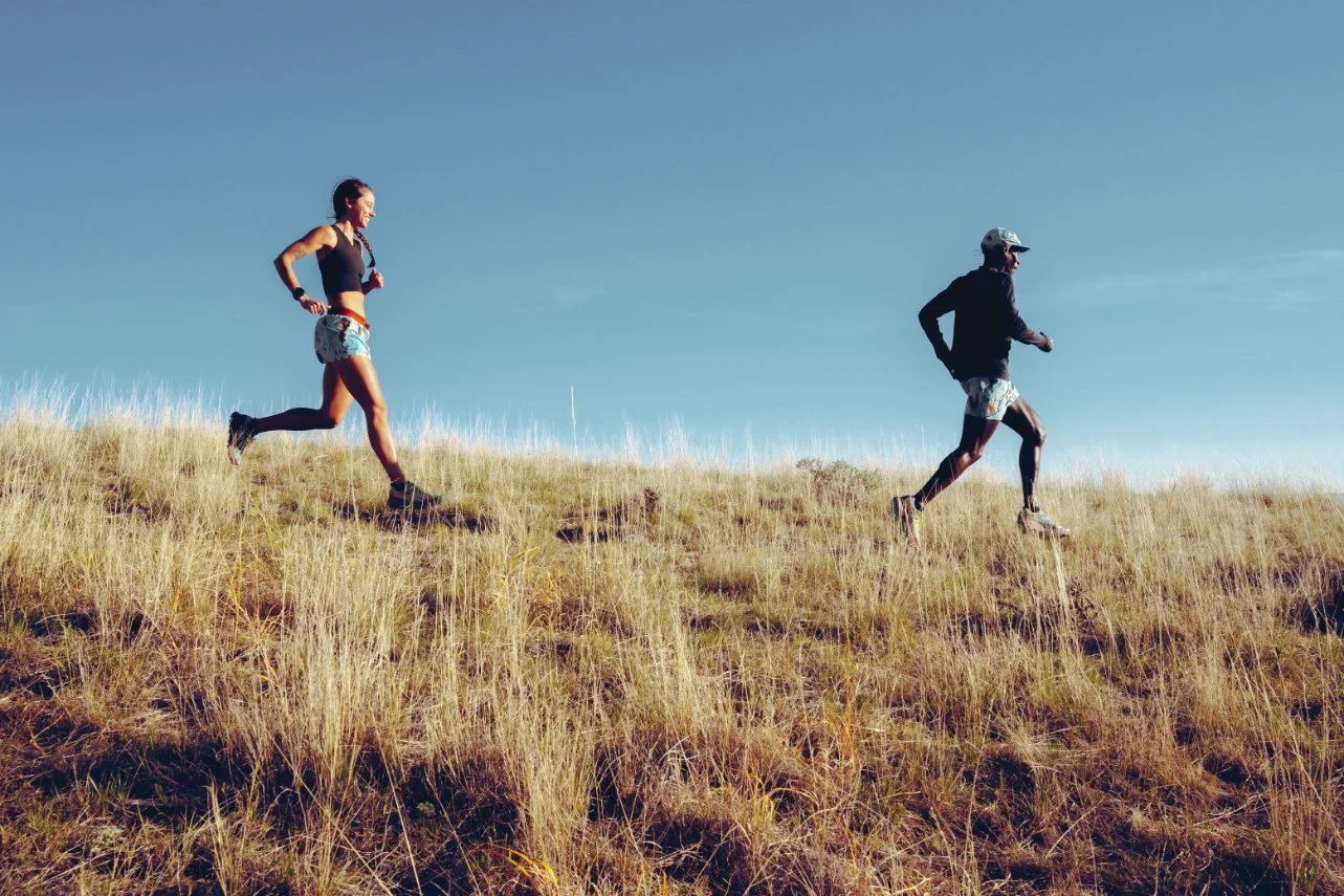 Two runners, a woman and a man, running uphill on a grassy hill with a clear blue sky.