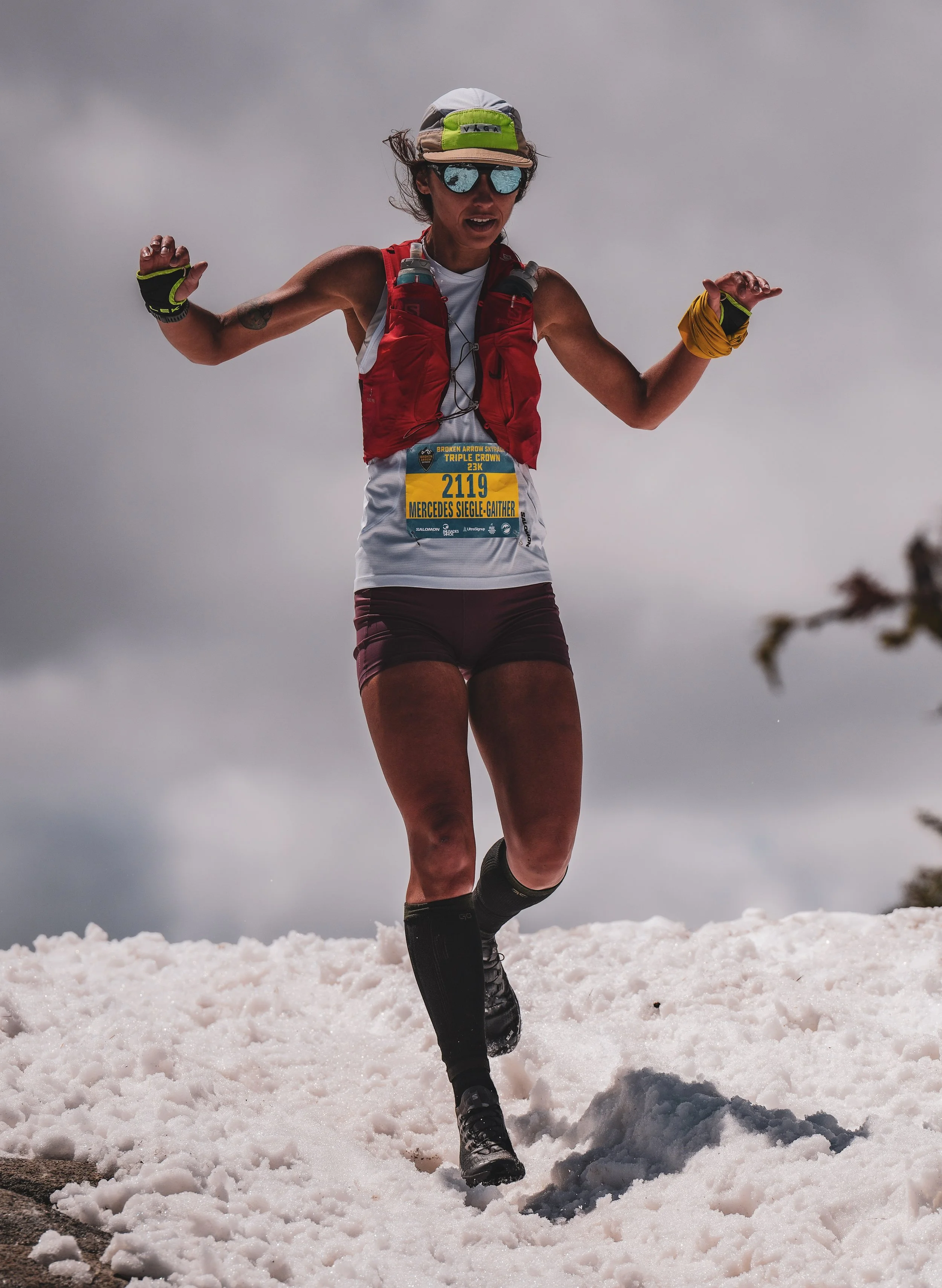 Female trail runner with race number 2119, wearing a red vest, white shirt, maroon shorts, black compression socks, and sunglasses, running on snow under cloudy sky.