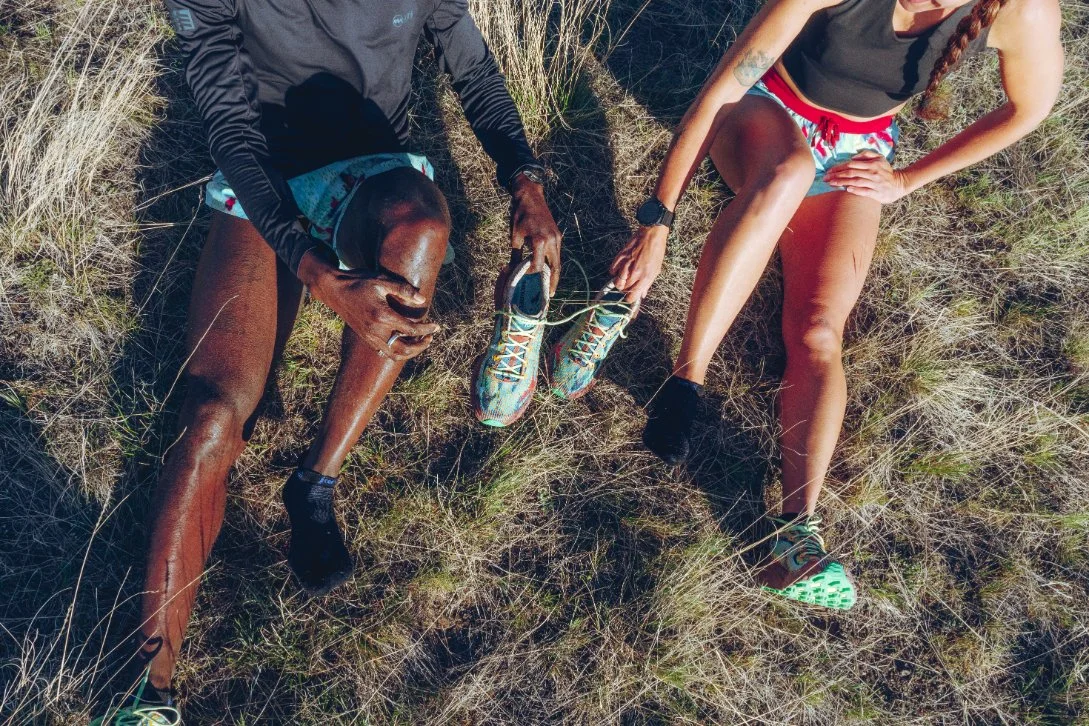 Two runners resting on a grassy outdoor trail, stretching and adjusting their shoes.