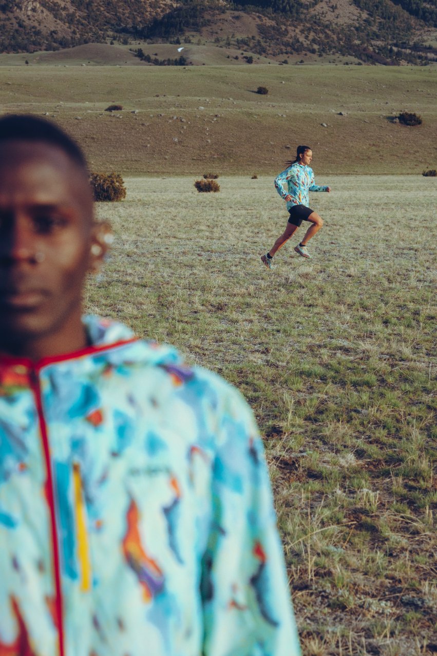 A blurry person in the foreground with earrings and colorful jacket, and another person running in the background in a large open field with hills.