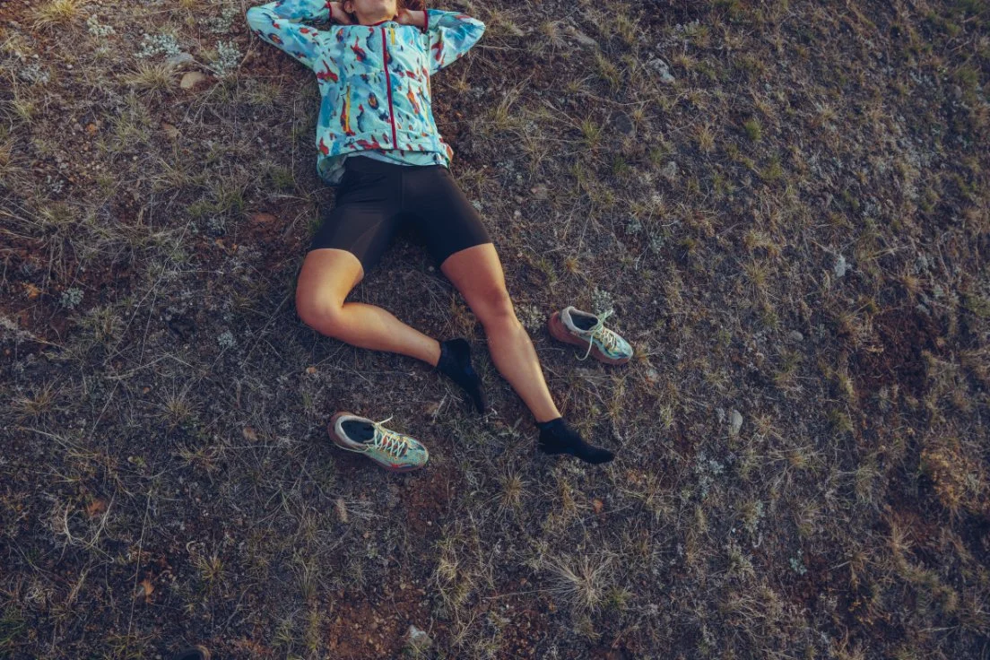 A person lying on their back on a grassy ground with hands behind their head, wearing a colorful jacket, black running shorts, and black socks, with a pair of running shoes nearby.