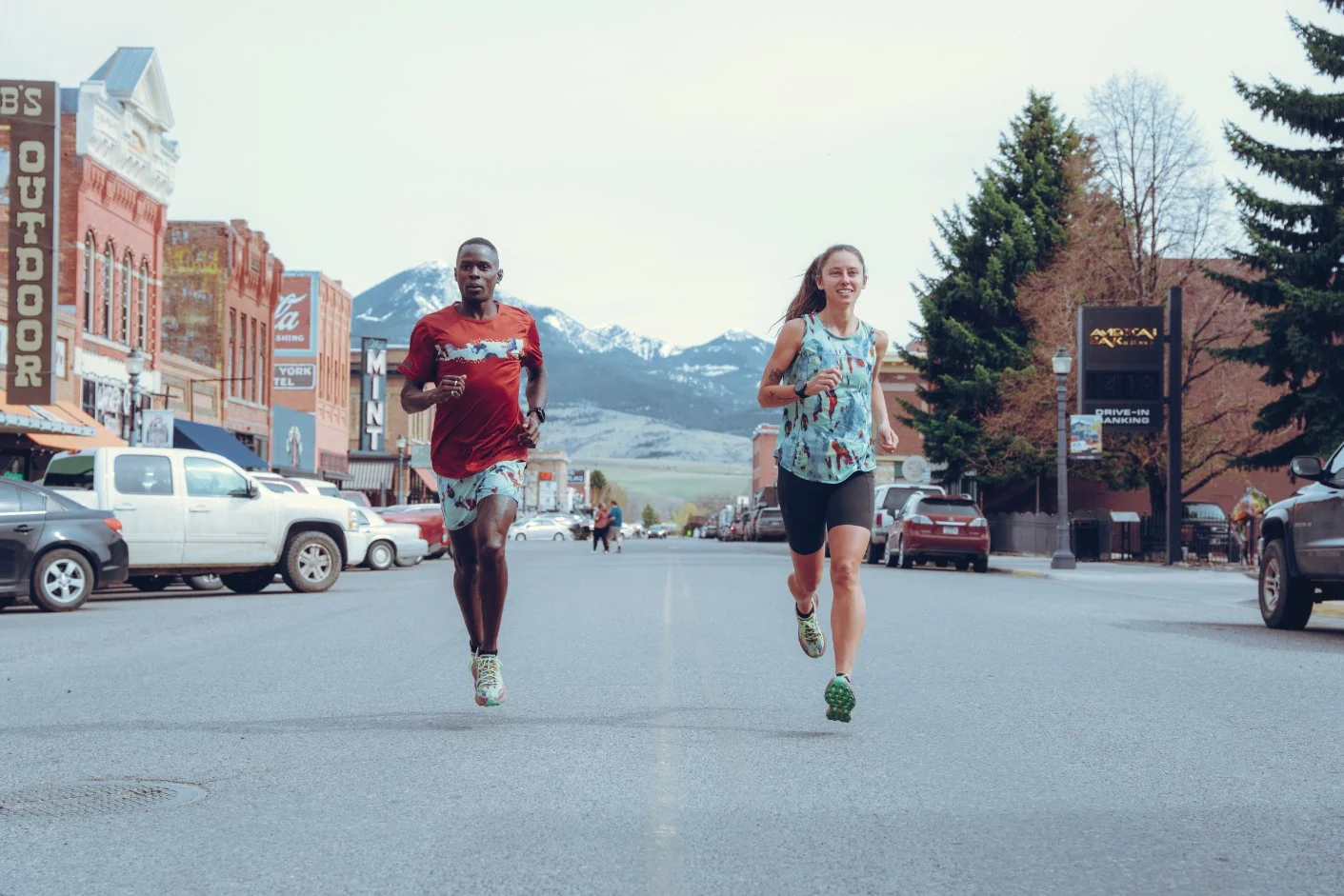 Two people running on a downtown street with mountains in the background, storefronts, parked cars, and trees on the sides.