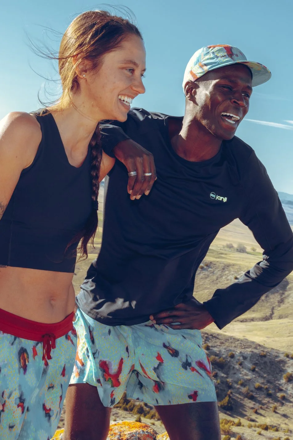 A young woman and man enjoying outdoors on a sunny day, smiling, with a scenic landscape of hills and clear blue sky in the background.