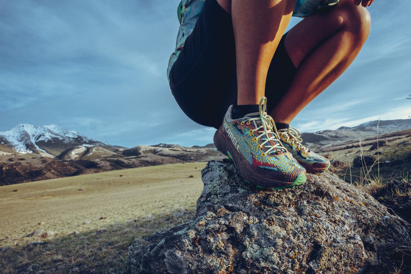 Person wearing colorful running shoes balancing on a rock in a mountainous outdoor landscape with snow-capped peaks in the background.