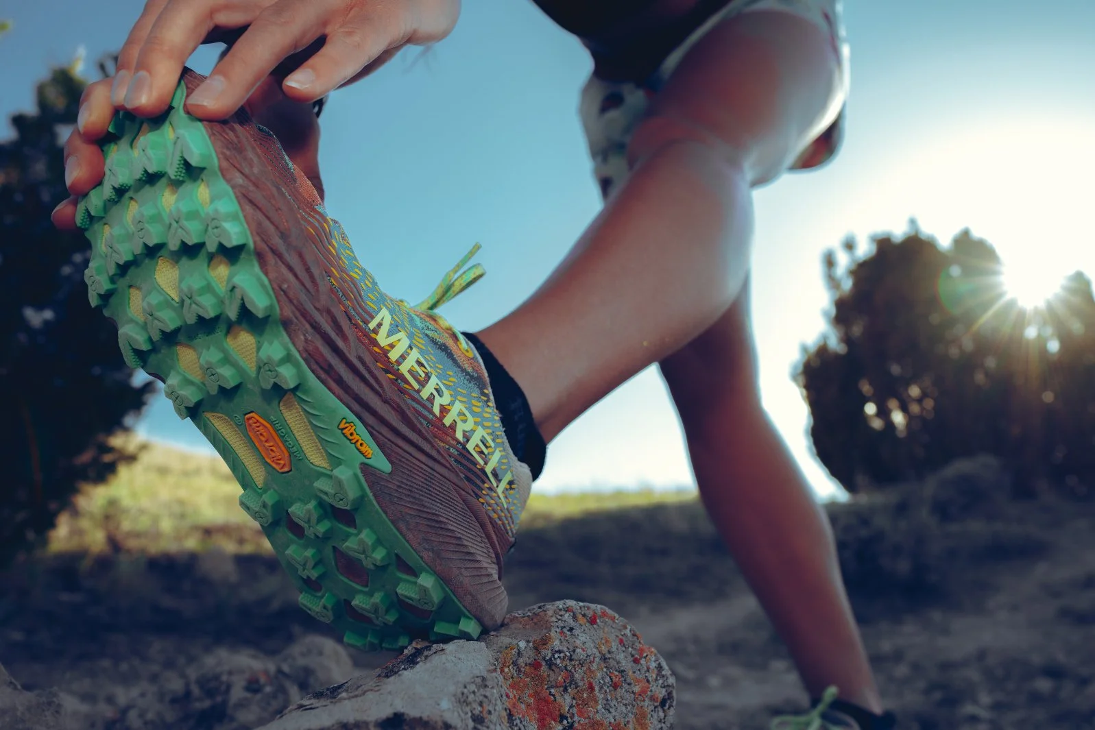 A person wearing brown and yellow trail running shoes with green soles is preparing to start running outdoors on a sunny day. The person is in a crawling position with their hand on the ground, and the sun is shining behind trees in the background.