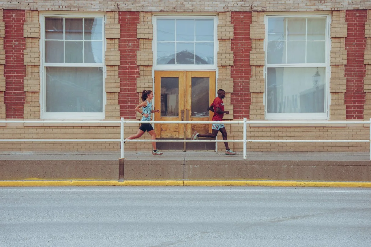 Two people running along a sidewalk in front of a brick building with large windows and a wooden door.