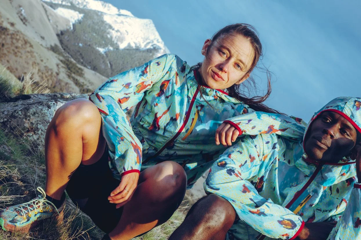 Two people in colorful outdoor jackets sitting on a rocky hillside with a canyon and blue sky in the background.