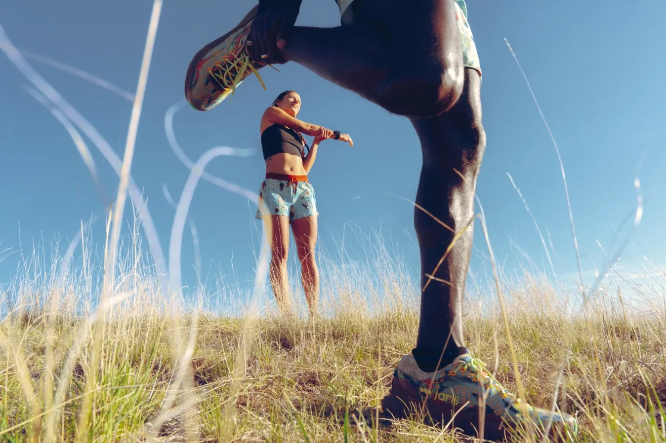 Low-angle view of a woman and a man on a grassy hill under a clear blue sky, the woman is stretching and the man is leaning forward, both dressed in athletic clothes and running shoes.