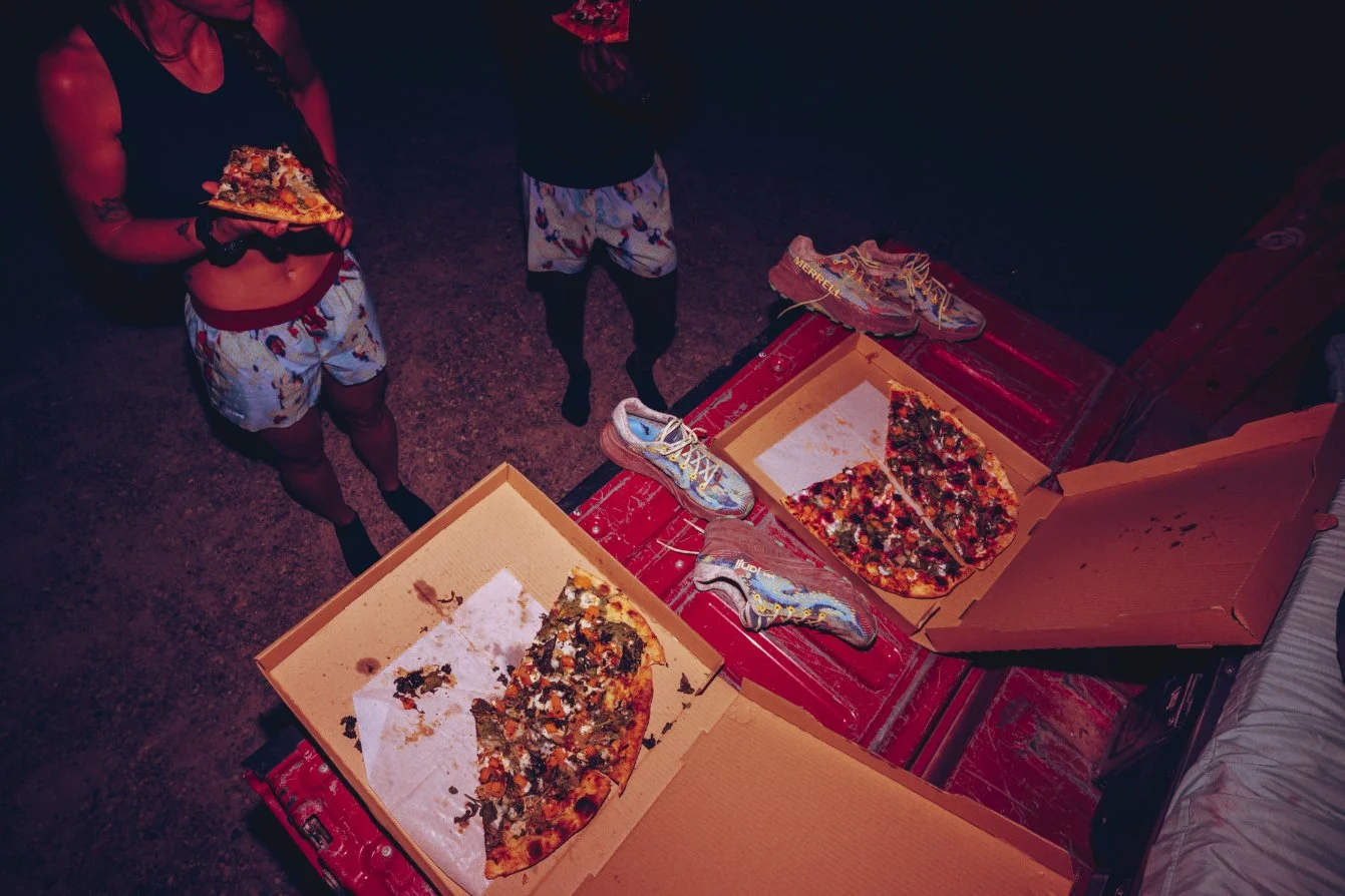 Two people standing around two open pizza boxes on a red surface, with leftover pizza slices and shoes nearby.