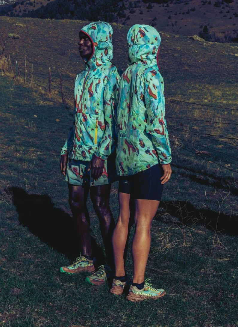 Two women wearing colorful, patterned outdoor jackets with hoods, standing back-to-back on a grassy hillside with mountains in the background.
