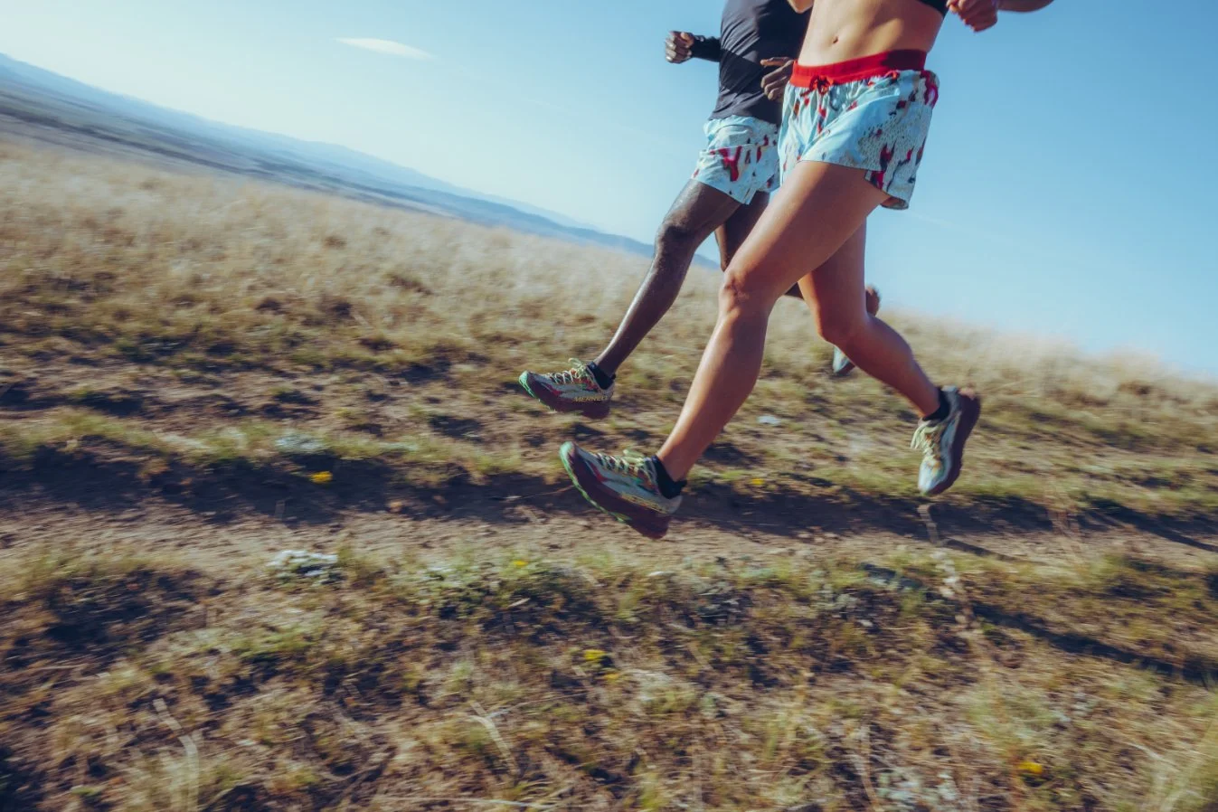 Two runners running outdoors on a grassy and slightly rocky trail in a sunny landscape.