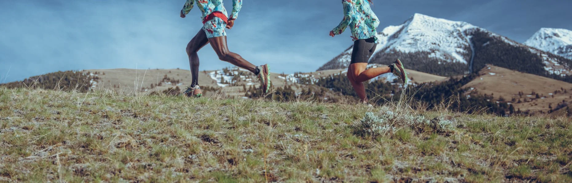 Two runners dressed in colorful outdoor clothing running on a grassy hillside with snow-capped mountains in the background.