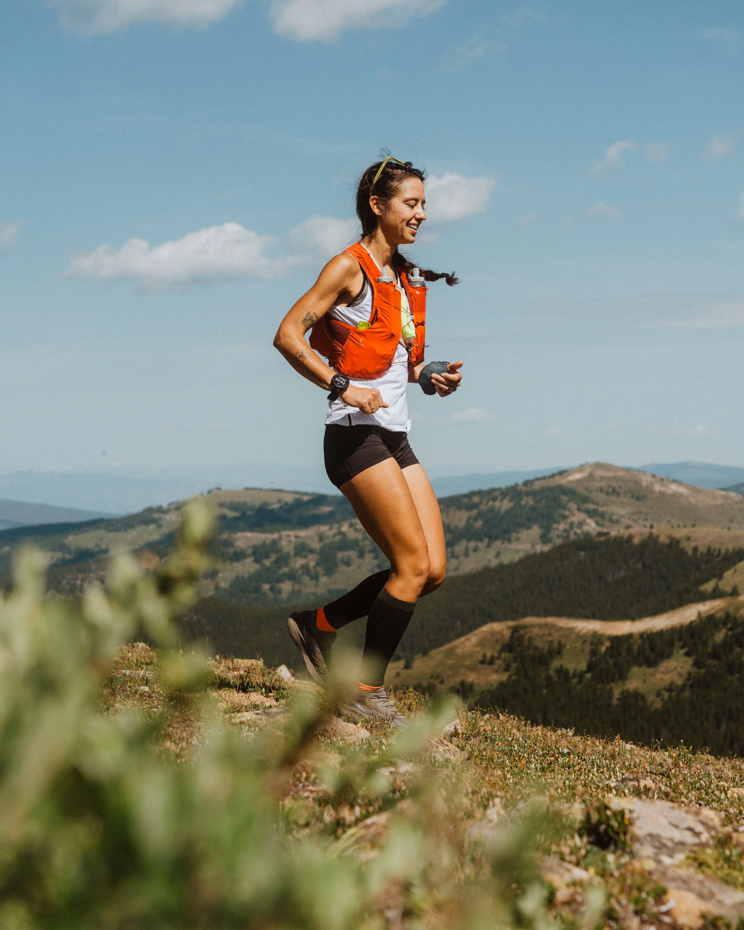 A woman trail running on a mountain trail with hills and blue sky in the background, wearing an orange hydration pack, black shorts, and running shoes.