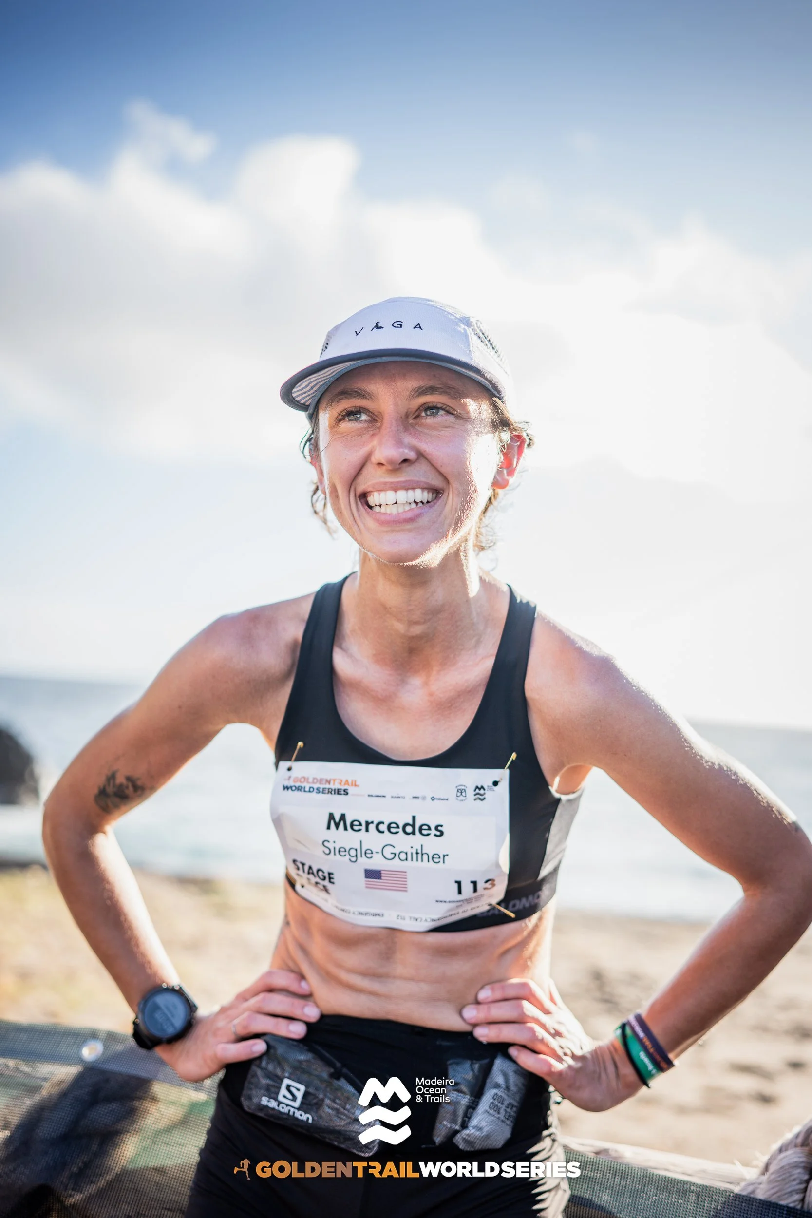 Smiling female marathon runner wearing a cap, race bib with name Mercedes and name tag Siegle-Gaither, standing on a beach with the ocean in the background.